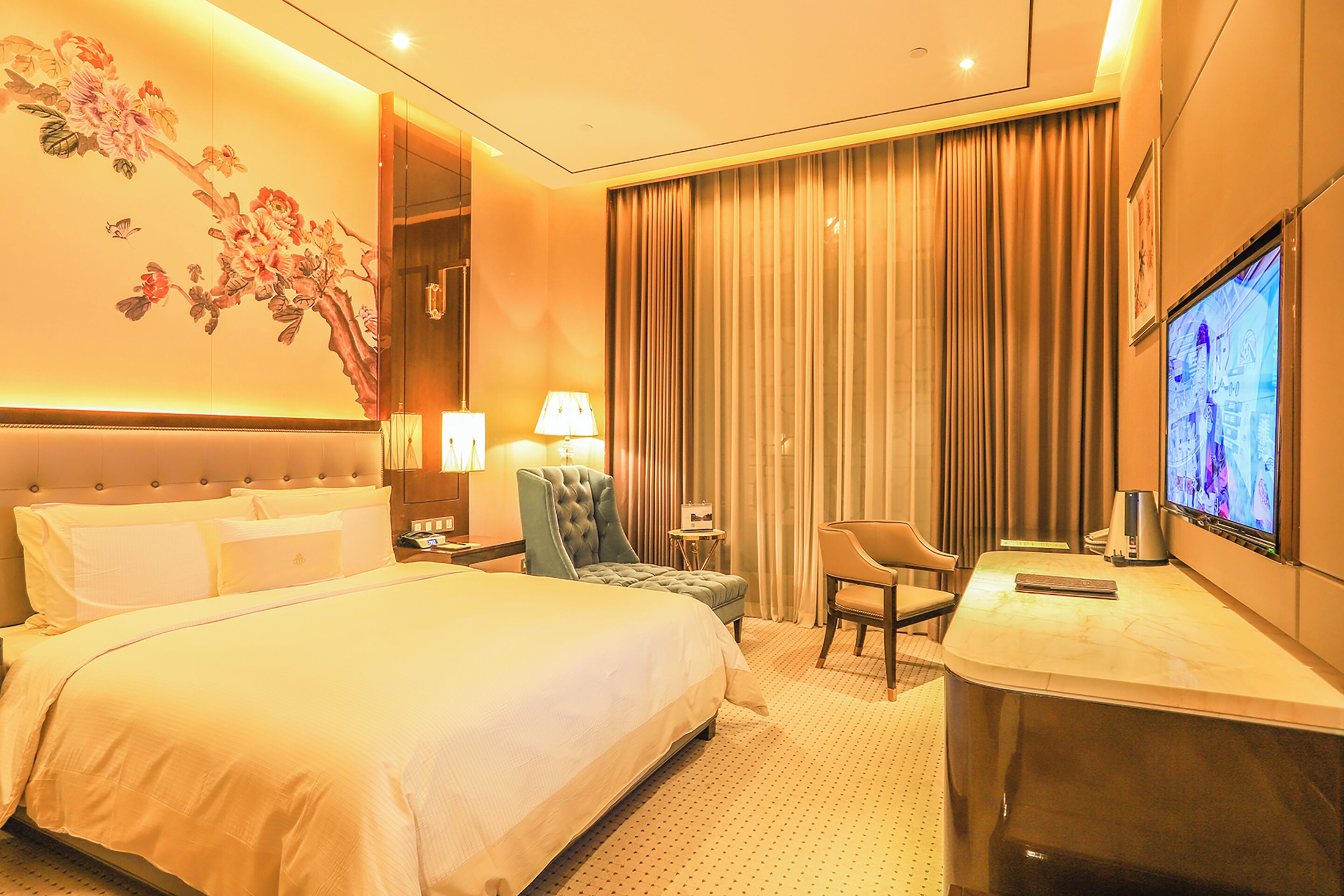 A hotel room with a large bed, a desk, a chair, a TV, and a wall with a floral mural.