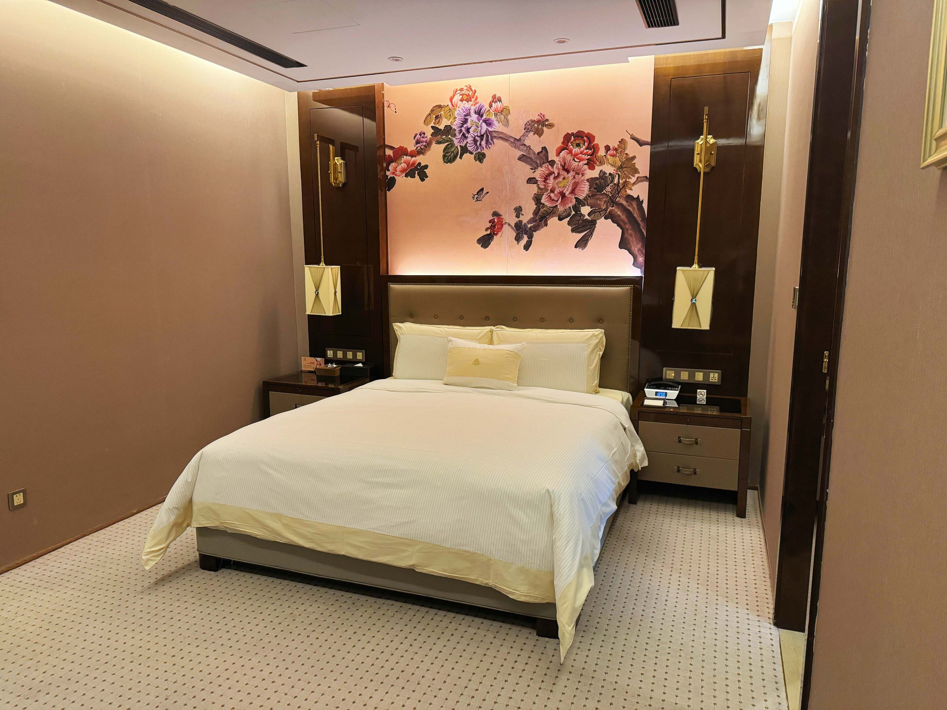 A hotel room with a large bed, a nightstand with a lamp, a wall-mounted artwork, and a door with a handle.