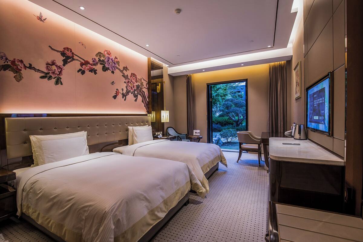Une chambre d’hôtel avec deux lits, un bureau, une télévision et une grande fenêtre donnant sur un paysage verdoyant.