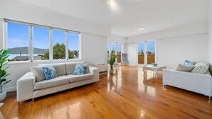Ein geräumiges Wohnzimmer mit Holzboden, zwei weißen Sofas und großen Fenstern mit Blick ins Freie.