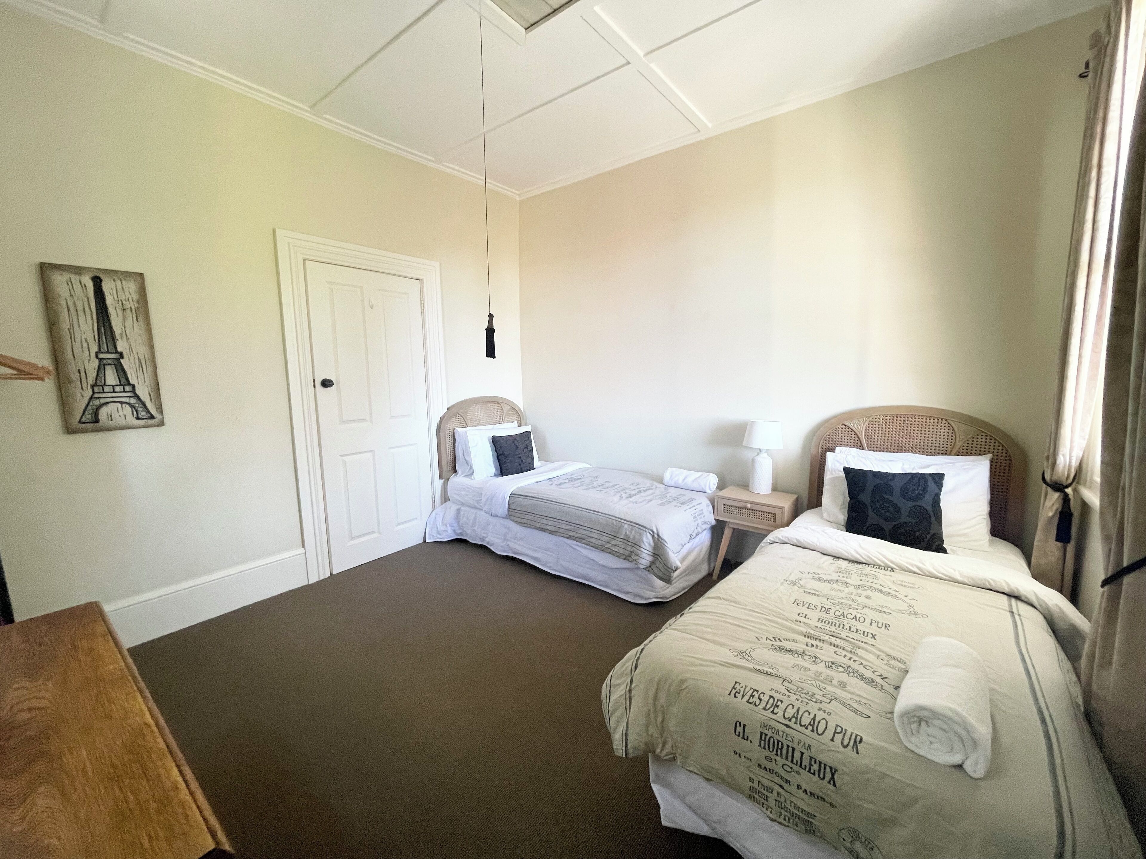 A hotel room with two beds, a door, a nightstand with a lamp, and a wall with a framed picture.