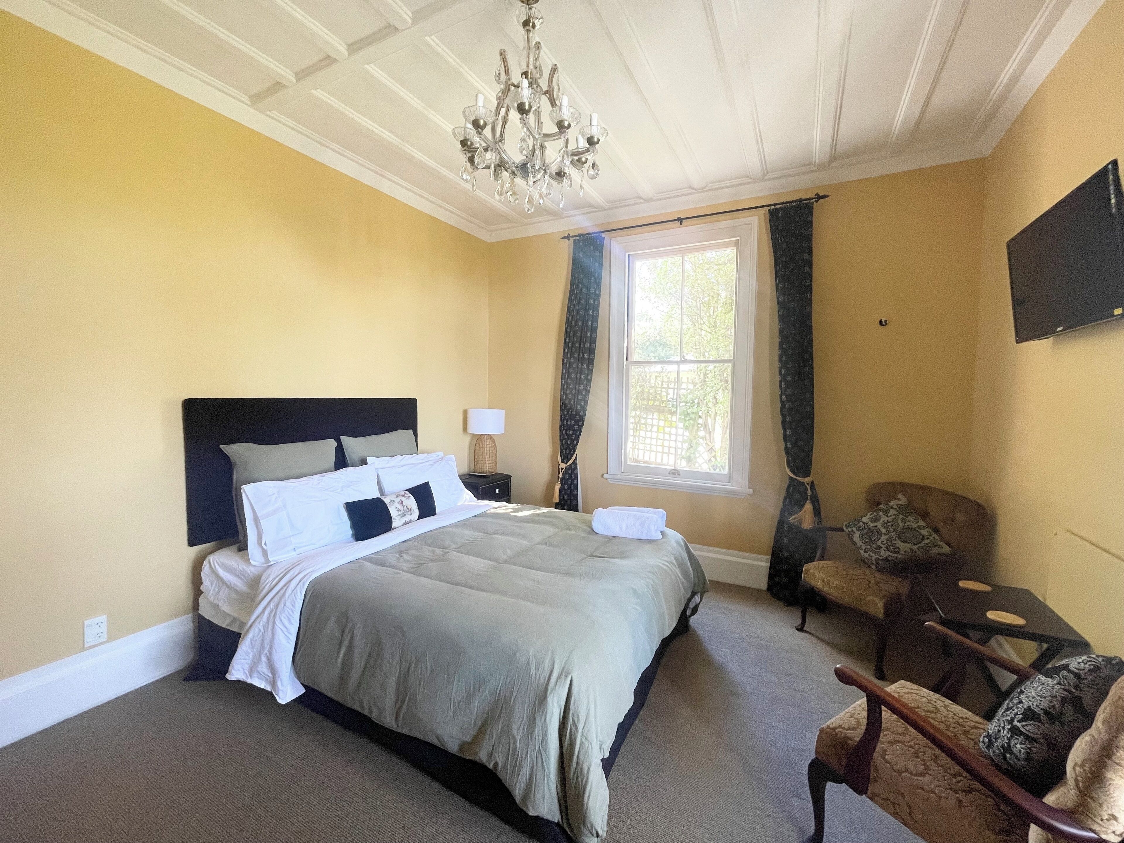 A bedroom with a bed, a nightstand, a lamp, a chair, a window, and a chandelier.