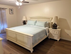 Une chambre à coucher comprenant un lit, des tables de chevet, un ventilateur de plafond et une fenêtre avec des rideaux.