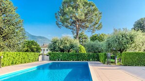 A rectangular swimming pool with clear blue water, surrounded by a paved patio and lush greenery, including a large tree and various shrubs. - La Breva by Interhome (Domaso)