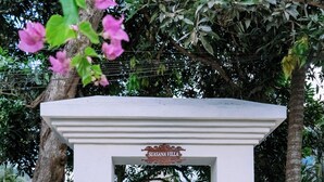 Une entrée blanche surmontée d’une structure à toit de chaume, flanquée de deux statues de lions en pierre, et portant une enseigne indiquant « SUASANA VILLA ».