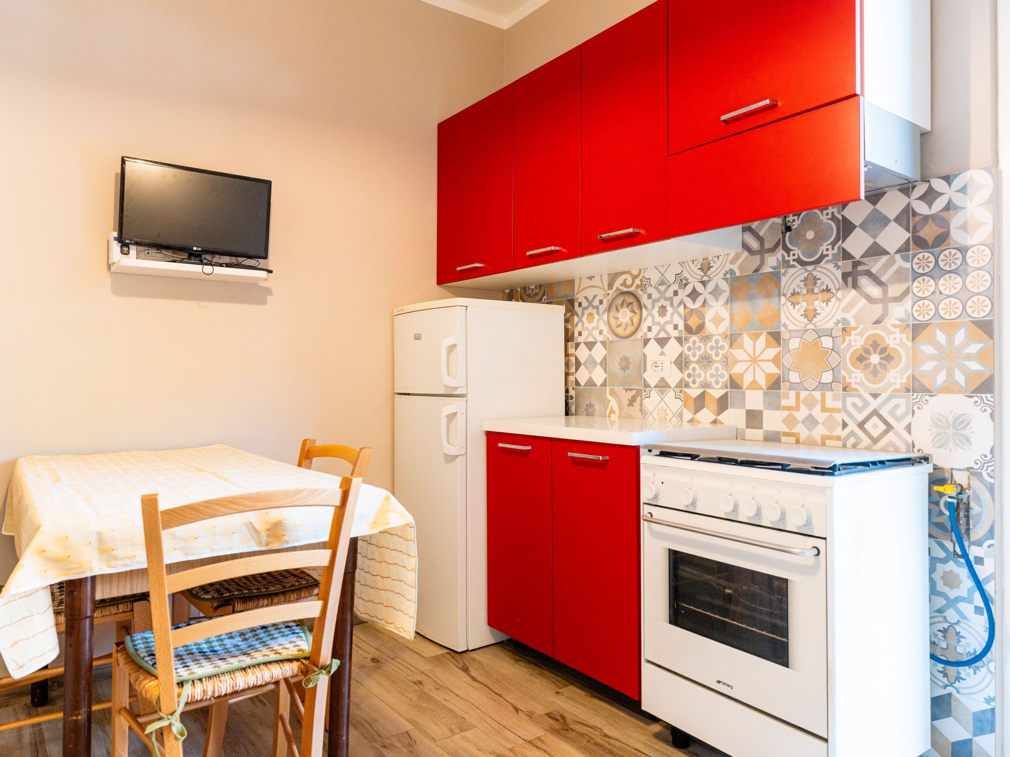 A compact kitchen with red cabinets, a white refrigerator, a stove, and a television mounted on the wall.
