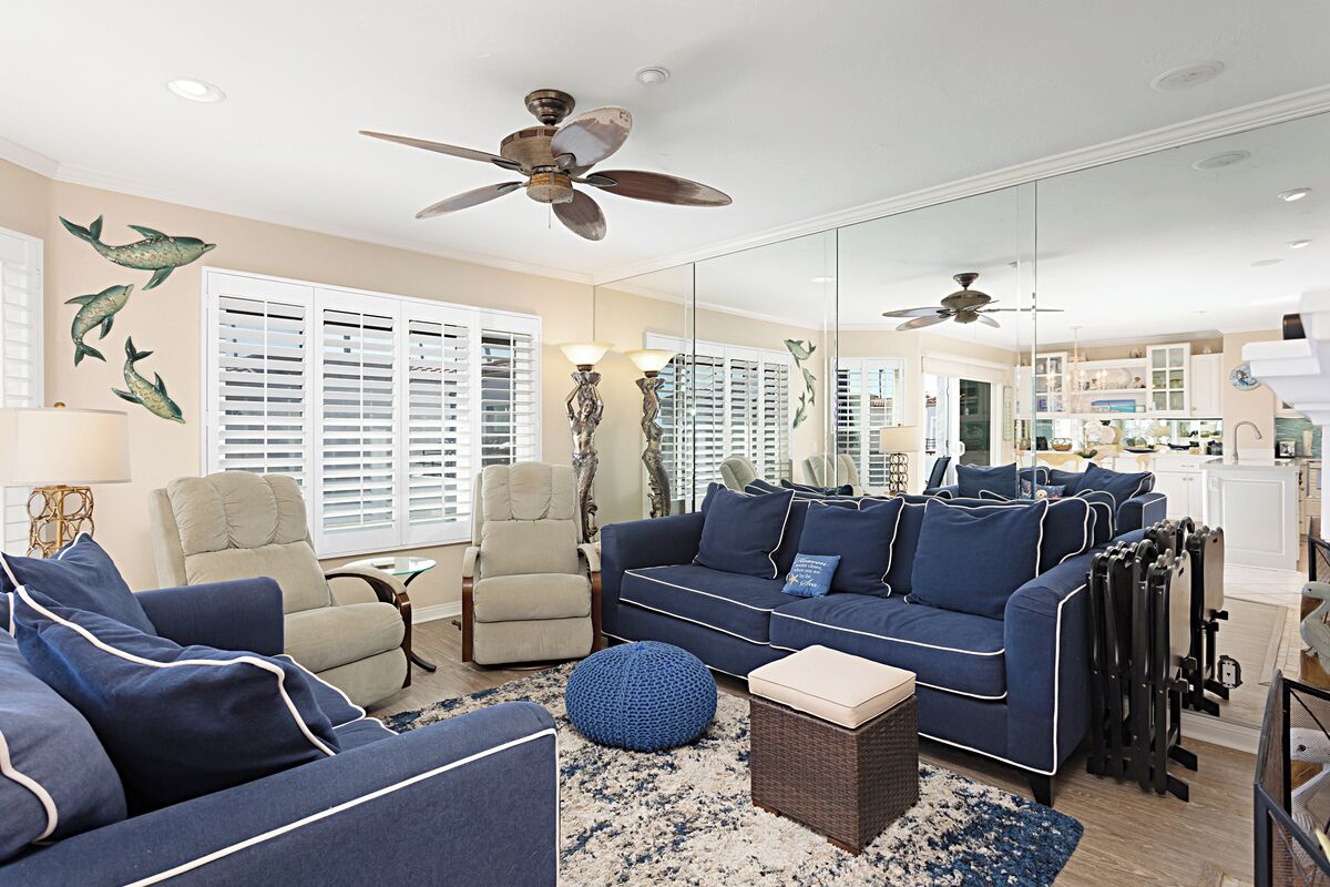 A living room with a blue sofa, beige armchair, and a small round table. There are ceiling fans, a large window with blinds, and a kitchen area visible through the glass wall.