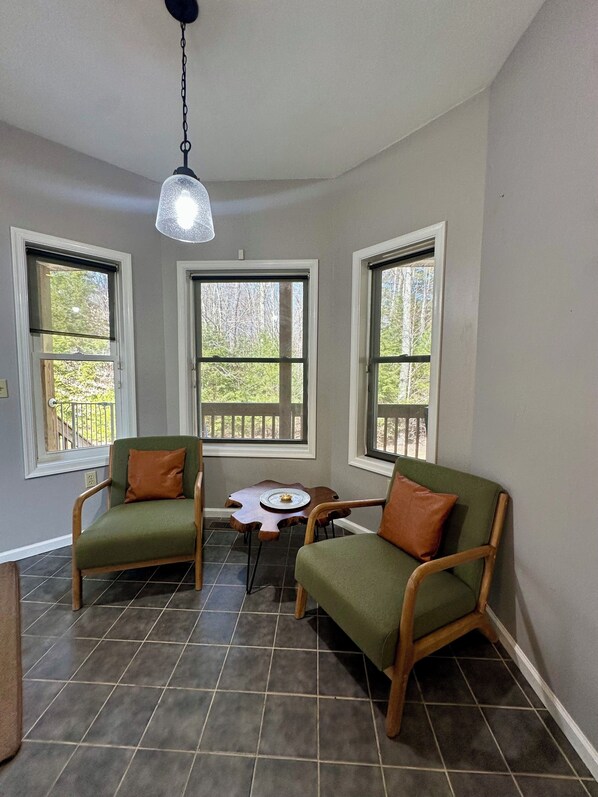 A room with two green armchairs, a small wooden table with a plate, and three windows with views of greenery. - Fire Pit, Hot Tub & Game Room Getaway In Pocono mountain (Blakeslee)