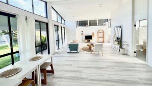 Uma sala de estar ampla com uma janela grande, uma mesa de jantar com bancos, um sofá e uma televisão ao fundo.