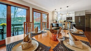 Un comedor con una mesa de madera preparada para una comida, una cocina al fondo y vistas a árboles a través de amplios ventanales.