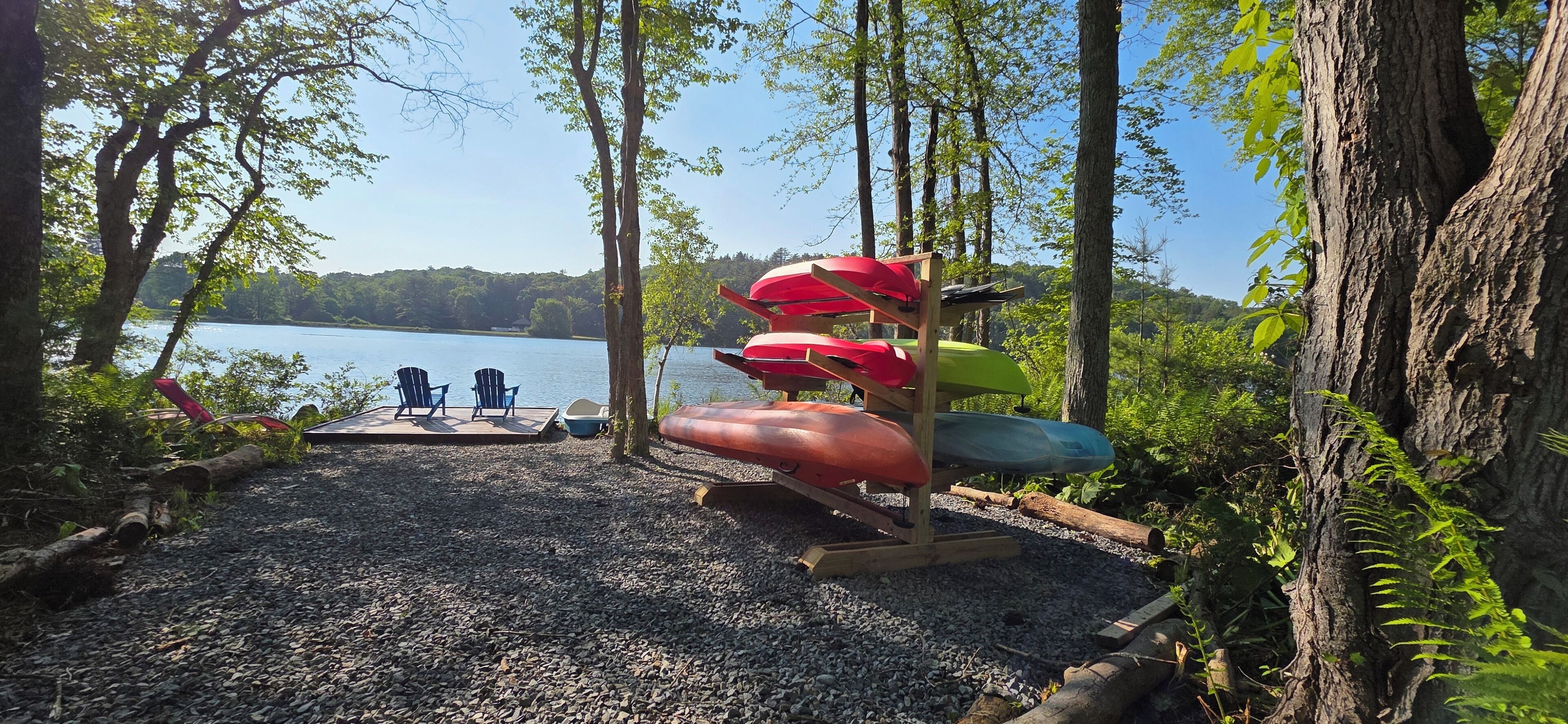 A lakeside setting with kayaks and paddleboards on a rack, surrounded by trees and a gravel path.