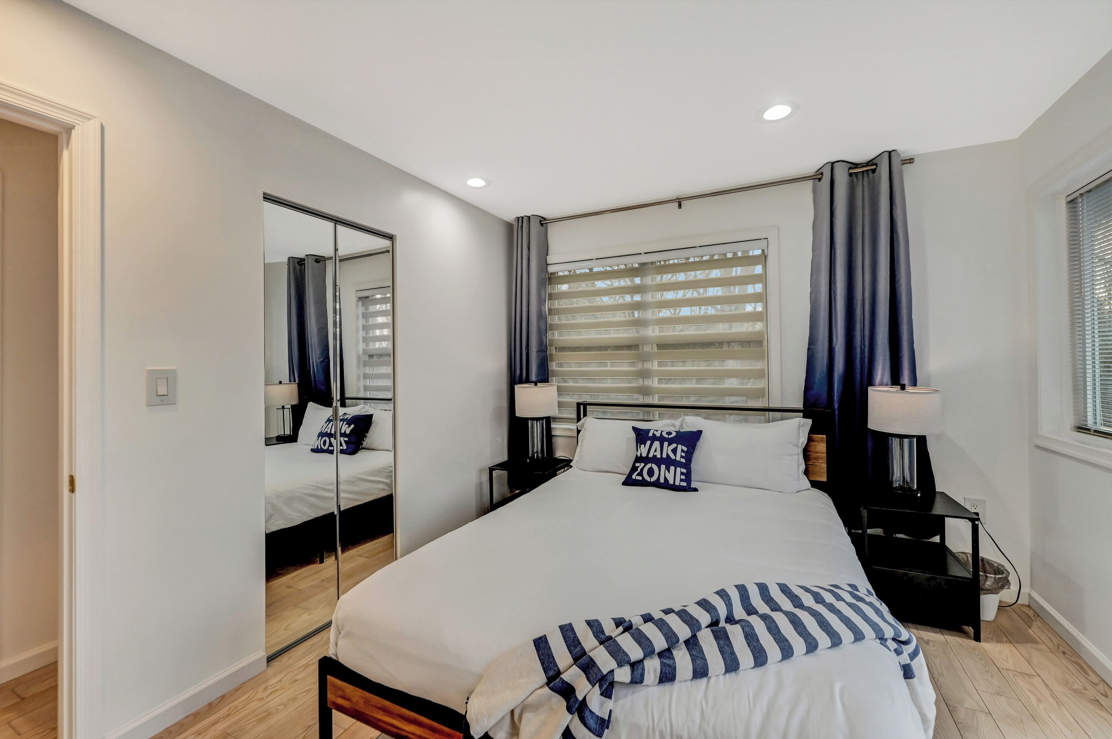 A modern bedroom with a large bed, a mirror, a bedside table, and a window with blinds.