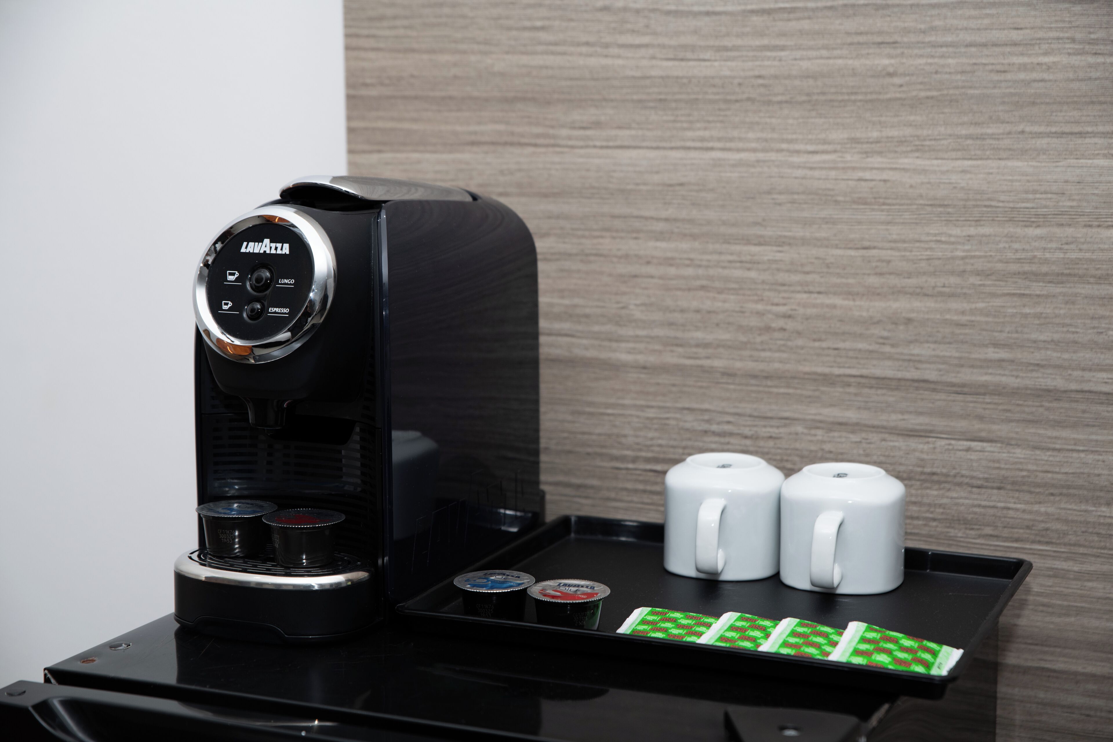 Una cafetera con un panel de control, dos tazas blancas y una bandeja con cápsulas de café.