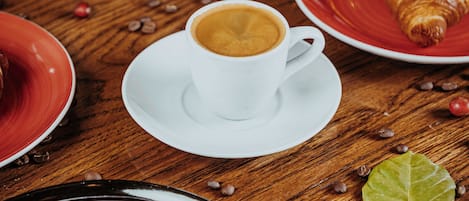 Una mesa de madera con una taza blanca de café, un plato rojo con un croissant y un plato negro con un pastelito a rayas rojas y blancas.