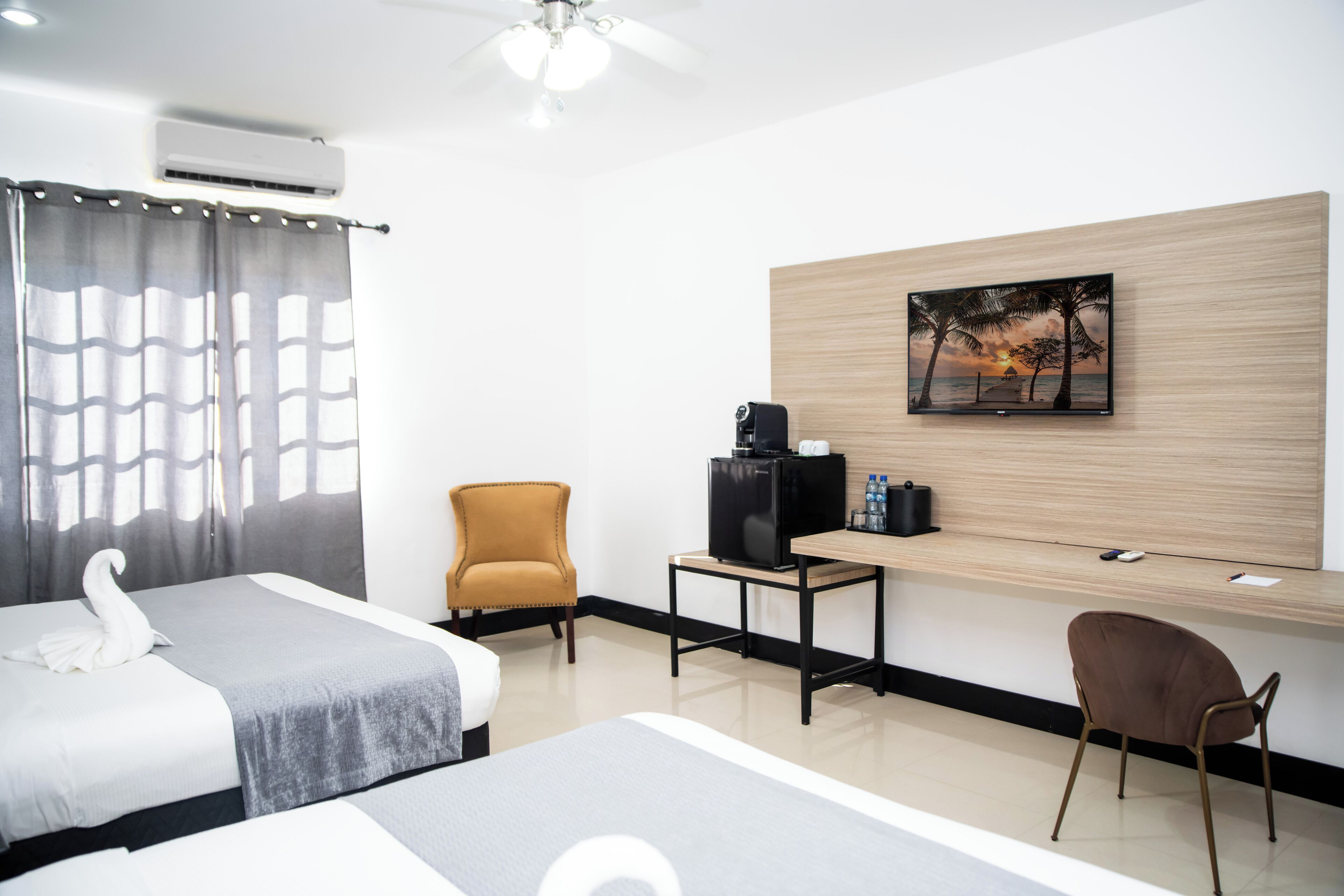 A hotel room with two beds, a desk with a computer and a coffee machine, a chair, and a wall-mounted TV.