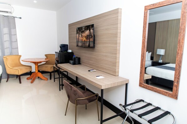 Une chambre d’hôtel avec un bureau en bois, deux chaises, une petite table, un lit avec une table de chevet, une lampe et un grand miroir.