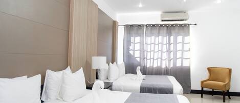 A hotel room with two beds, a ceiling fan, an air conditioning unit, a lamp, a chair, and a window with curtains.
