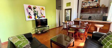 A living room with a green wall, a black leather sofa, a glass coffee table, and a TV on a stand.
