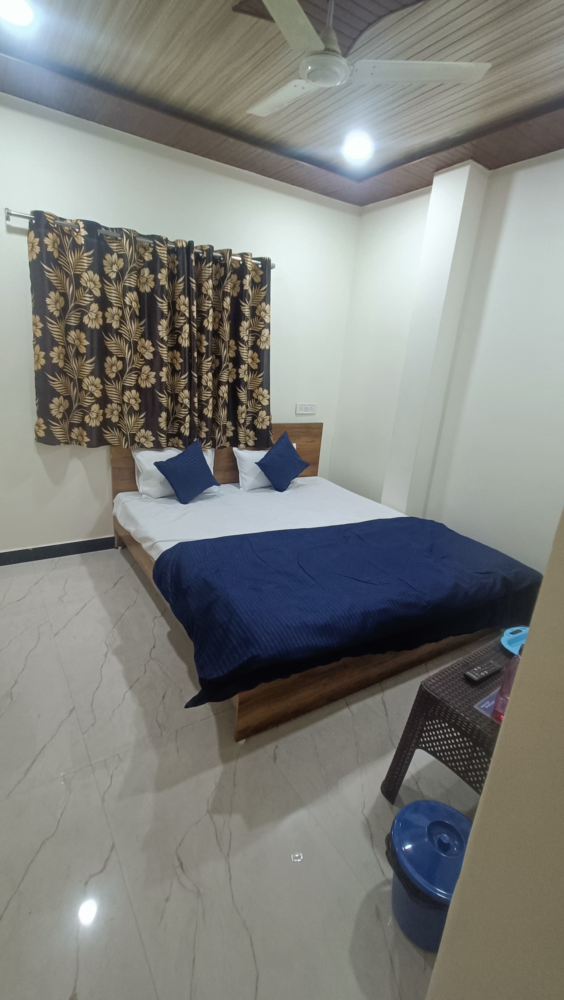 A bedroom with a bed, a ceiling fan, a window with floral curtains, and a small table with a blue container.