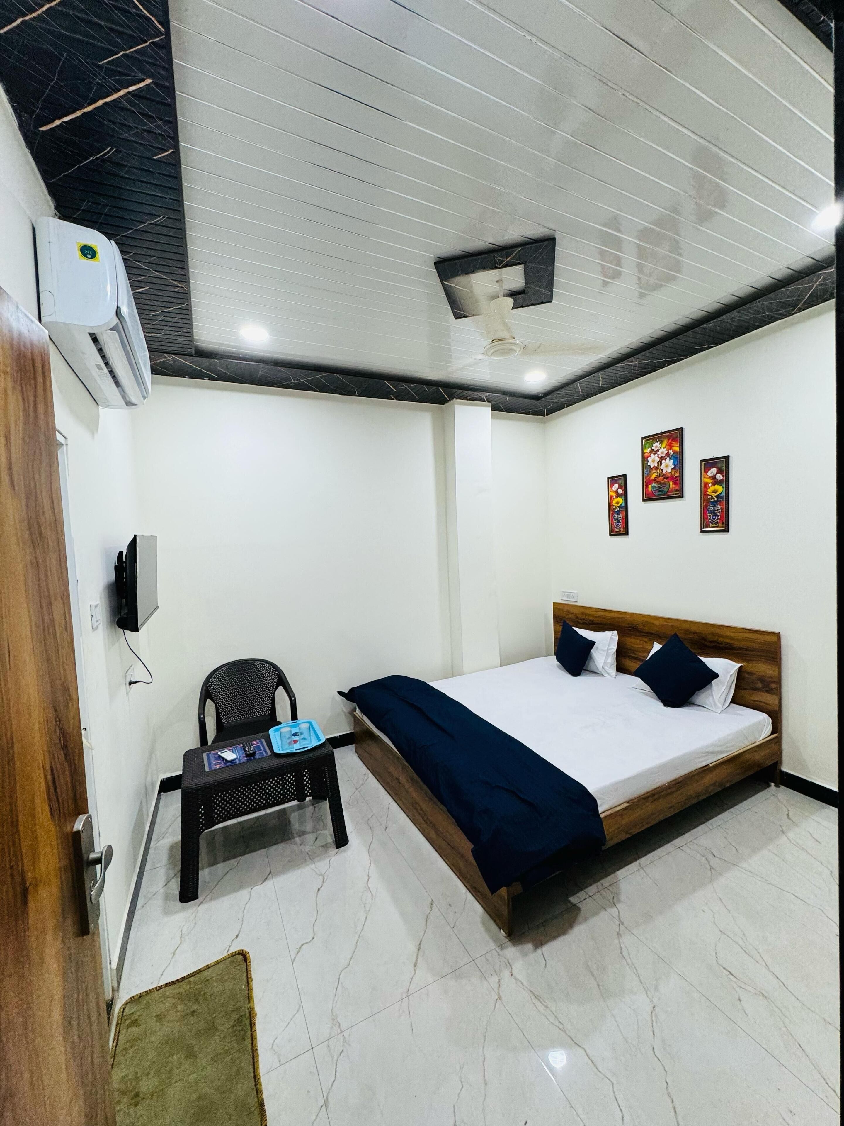 A bedroom with a bed, a chair, a small table, an air conditioning unit, and three wall-mounted paintings.