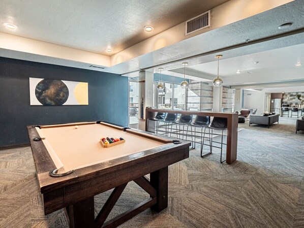 Une table de billard dans une pièce dont le sol est recouvert d’un tapis à motifs chevrons, un coin salon avec des fauteuils et une grande fenêtre laissant entrer la lumière naturelle.