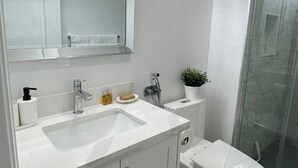 Un baño moderno con lavamanos, espejo y inodoro blancos. Incluye zona de ducha con puerta de cristal y una pequeña planta en el mostrador.
