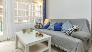 A modern living room with a grey sofa, blue and patterned cushions, a white coffee table with a plate of food, and a large window with a view of a building.