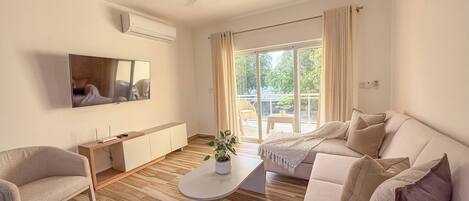 Una sala de estar moderna con sofá, sillón y una mesa de centro. Hay un televisor de pantalla plana montado en la pared, un ventilador de techo y una puerta corrediza de vidrio que da a un área exterior.