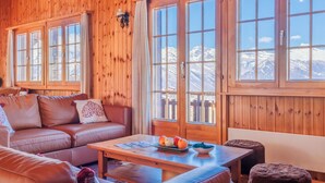 Una habitación acogedora con paredes de madera, un sofá de cuero, una mesa de madera con fruta y grandes ventanales con vistas a la montaña.