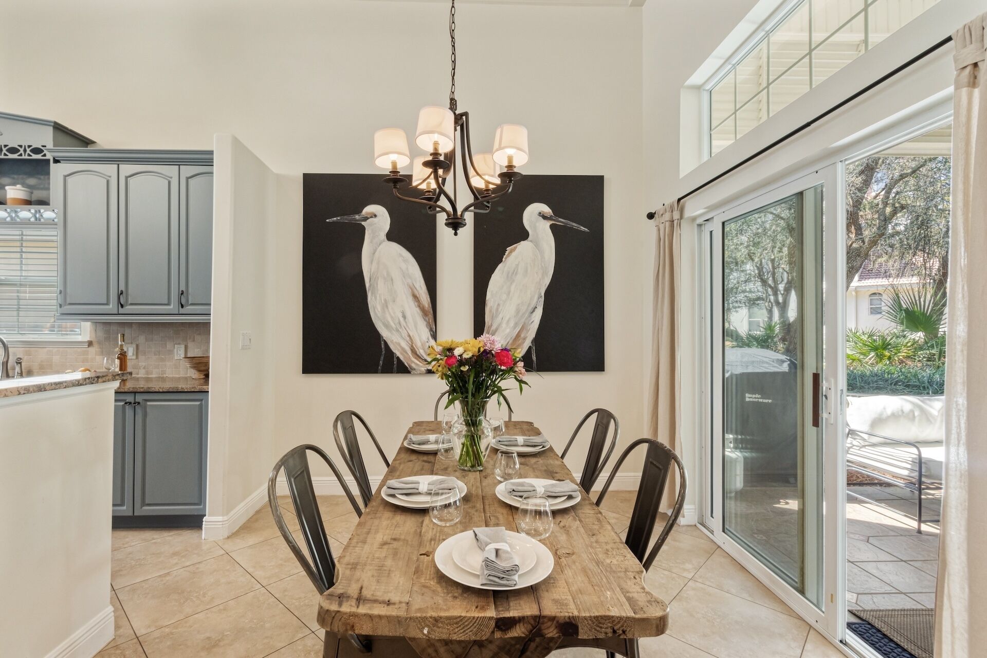 Un coin repas avec une table en bois dressée pour six personnes, un lustre et un grand tableau représentant un oiseau accroché au mur.