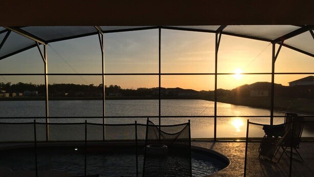 Een overdekt buitengebied met een zwembad, zitplaatsen en uitzicht op een zonsondergang over het water.