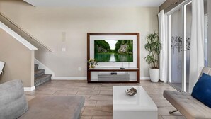 A modern living room with a flat-screen TV mounted on a wooden stand, a grey sofa, a white coffee table, and a staircase leading to another level.