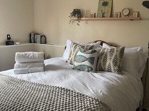 A neatly made bed with white linens and decorative pillows, a bedside shelf with books and a clock, and a bedside plant.