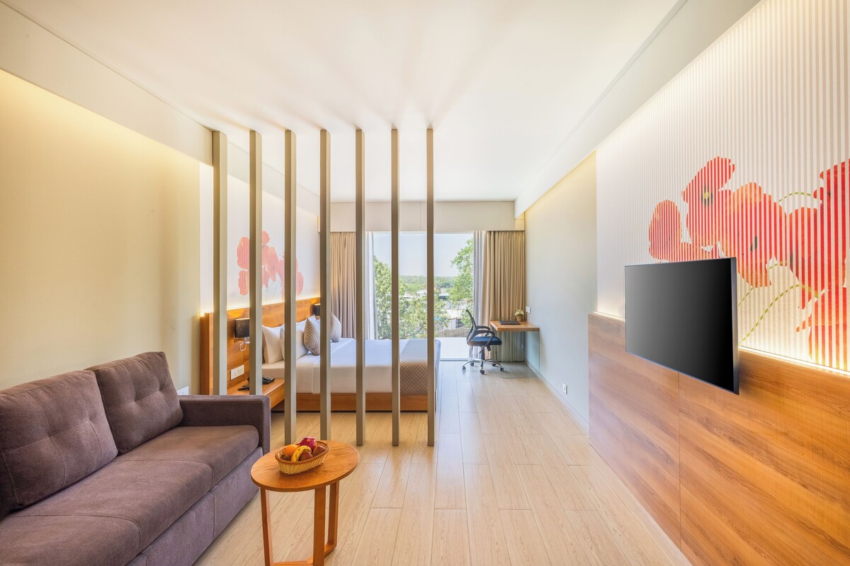 Habitación de hotel moderna con sofá, una pequeña mesa redonda con una cesta, televisor montado en la pared, escritorio con silla y un ventanal con vistas a zonas verdes.