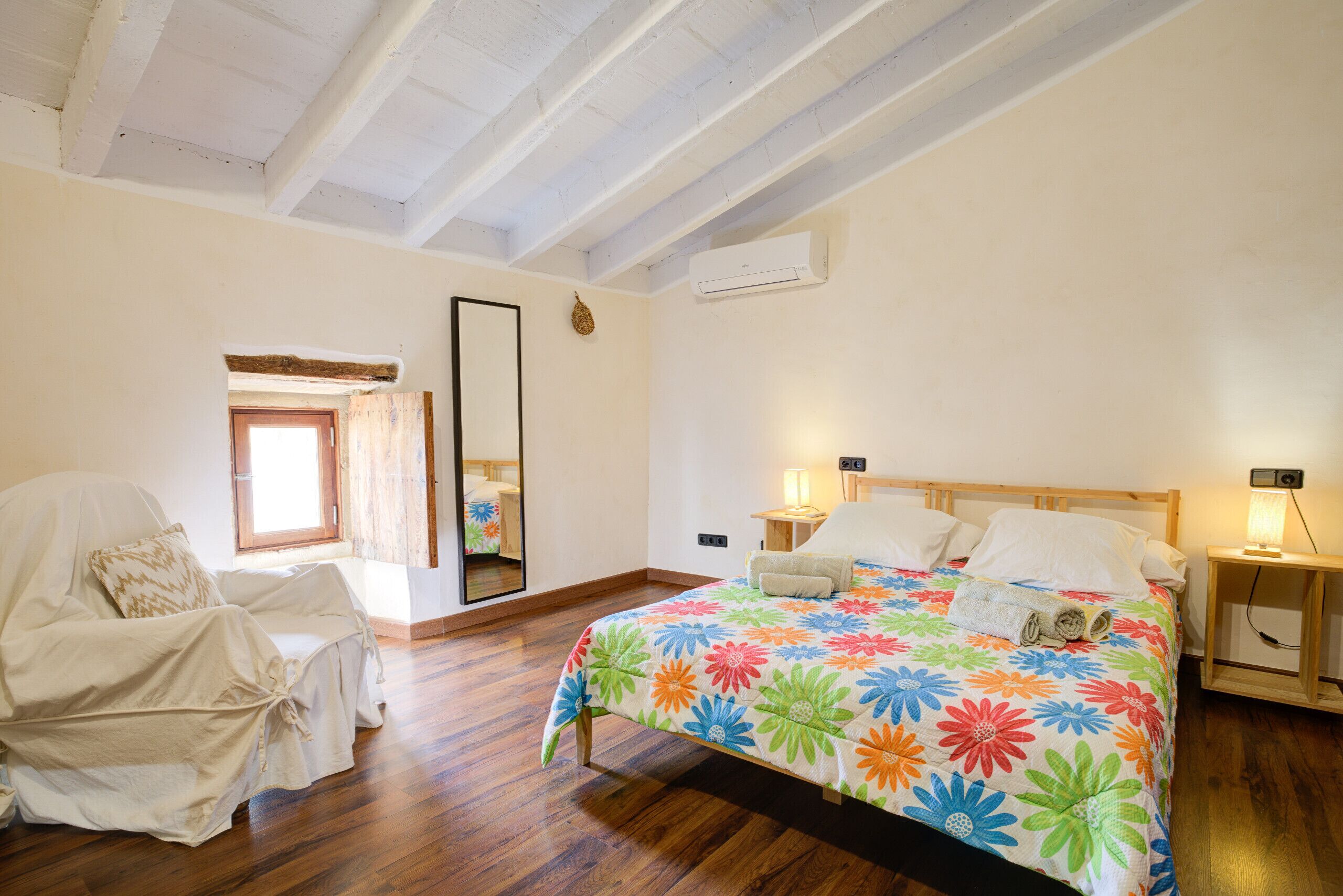A bedroom with a wooden floor, a bed with a floral bedspread, a chair covered in white, a nightstand with a lamp, and a mirror on the wall.