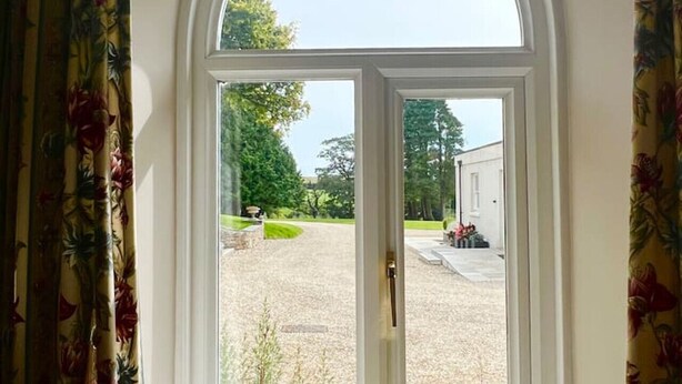 A window with a white frame and a view of a gravel driveway leading to a white building. - Coach House Quiet Country Retreat for 6 Near Coast (Loddiswell)