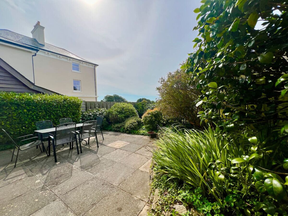 Une terrasse avec une table et des chaises, entourée d’une végétation luxuriante, avec une maison en arrière-plan.