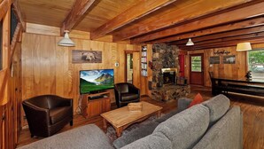 A cozy cabin interior with wooden walls, a stone fireplace, a television, leather chairs, and a sofa. - Sunshine Cabin Cozy 2-Story Getaway (Big Bear Lake)