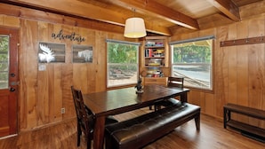 A wooden cabin interior with a dining table, benches, a bookshelf, and large windows. - Sunshine Cabin Cozy 2-Story Getaway (Big Bear Lake)