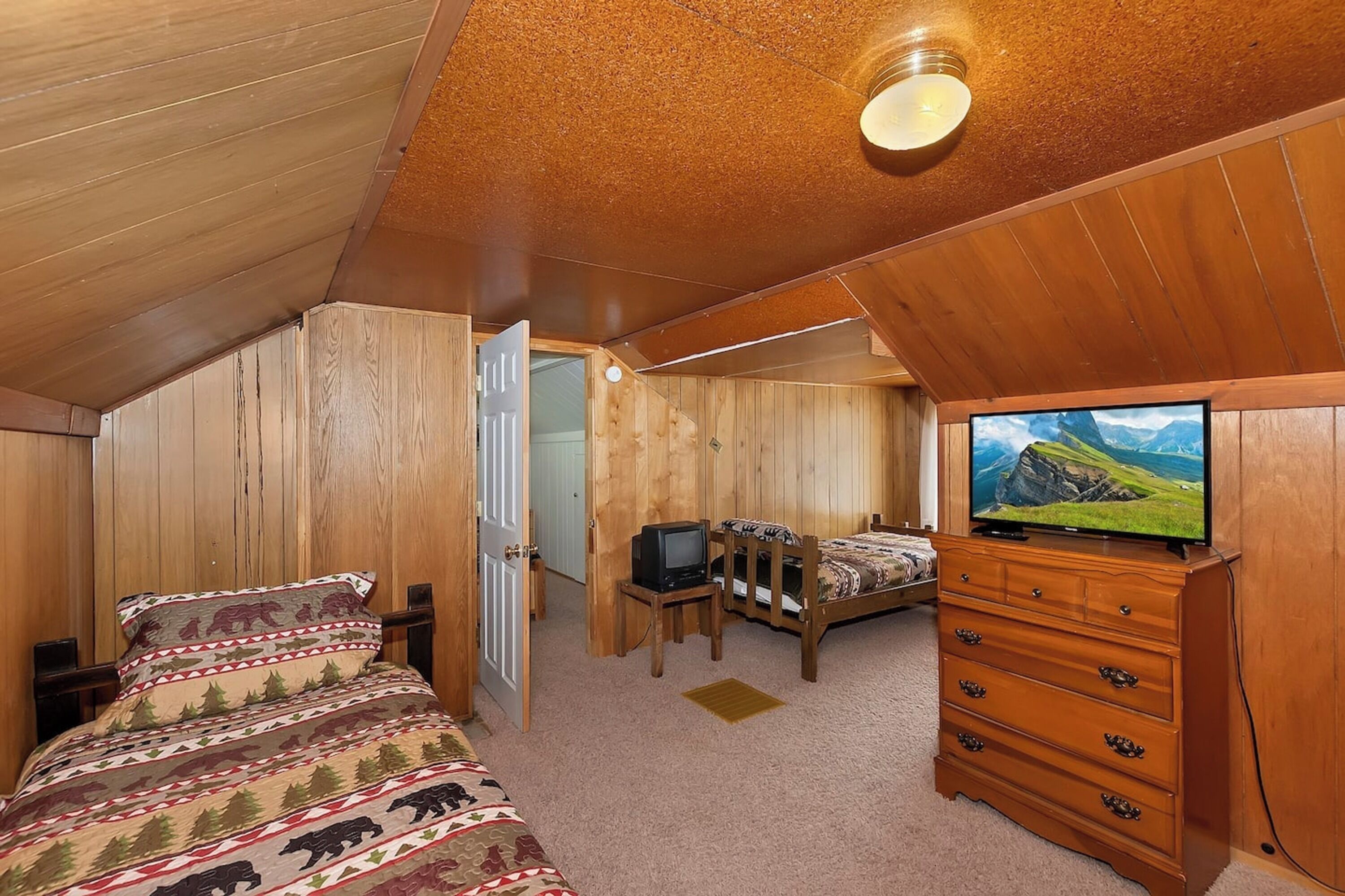 Ein Zimmer mit Holzwänden, einem Bett, einem Schminktisch, einem Fernseher und einem Schreibtisch mit Computer.