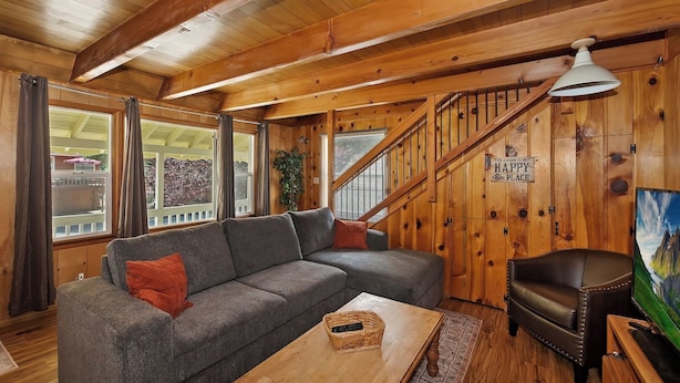A cozy cabin interior with a wooden staircase, a gray sectional sofa, a leather armchair, and a television on a wooden stand. - Sunshine Cabin Cozy 2-Story Getaway (Big Bear Lake)