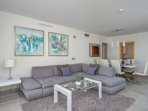 Ein modernes Wohnzimmer mit einer grauen Eckcouch, einem Glastisch, abstrakten Gemälden, einem Essbereich mit weißem Tisch und Stühlen sowie einer im Hintergrund sichtbaren Küche.