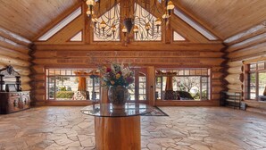 A spacious log cabin interior with a stone floor, a central glass-top table, and a chandelier. - Legacy Lodge & Barn Retreat, Sleeps 42 (Heber)