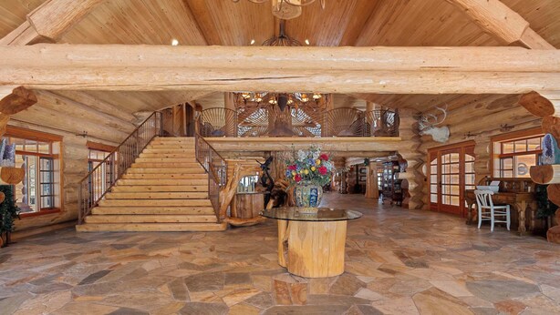 A spacious log cabin interior with a stone-tiled floor, a central round table, and a staircase leading to an upper level. - Legacy Lodge & Barn Retreat, Sleeps 42 (Heber)