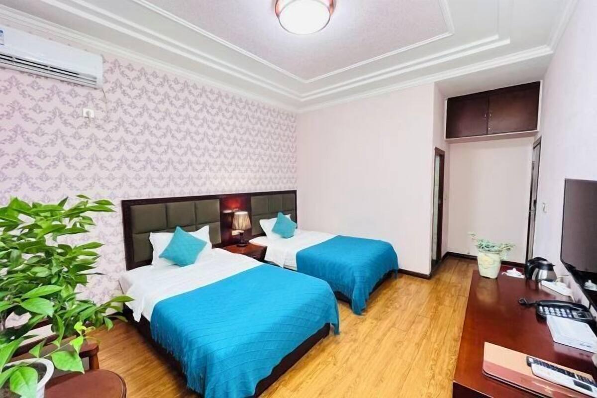 A hotel room with two beds, a desk, a chair, a plant, and an air conditioning unit.