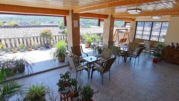 A covered outdoor seating area with tables and chairs, surrounded by potted plants, and overlooking a traditional tiled roof structure.