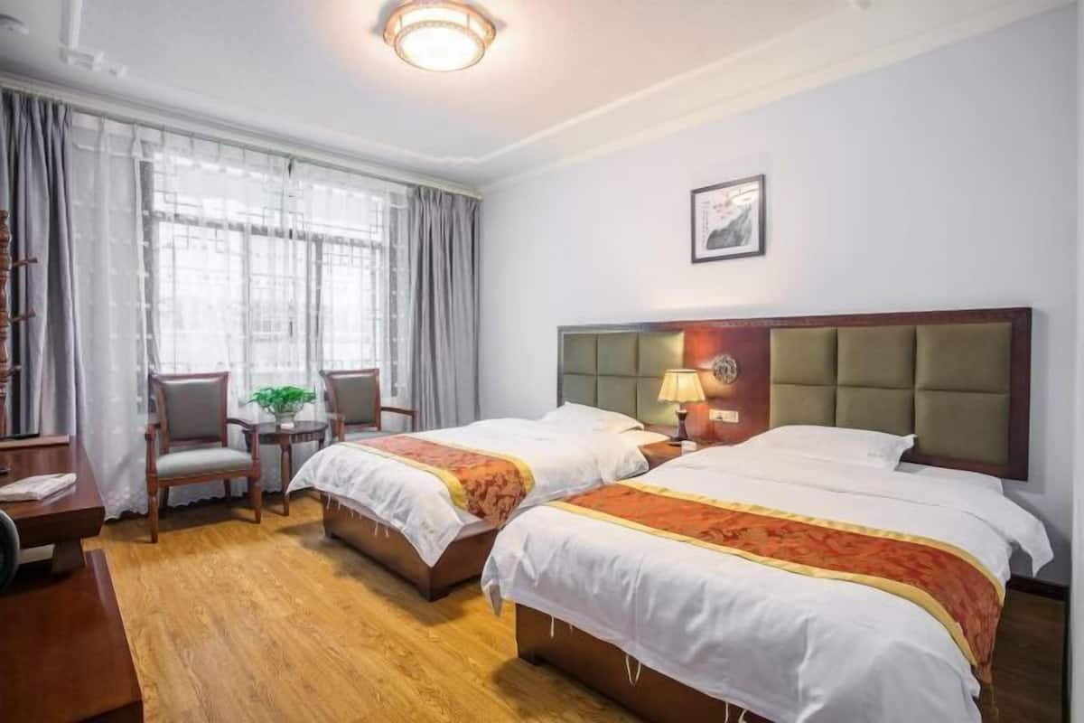 A hotel room with two beds, a desk, a chair, a small table with a plant, and a window with curtains.