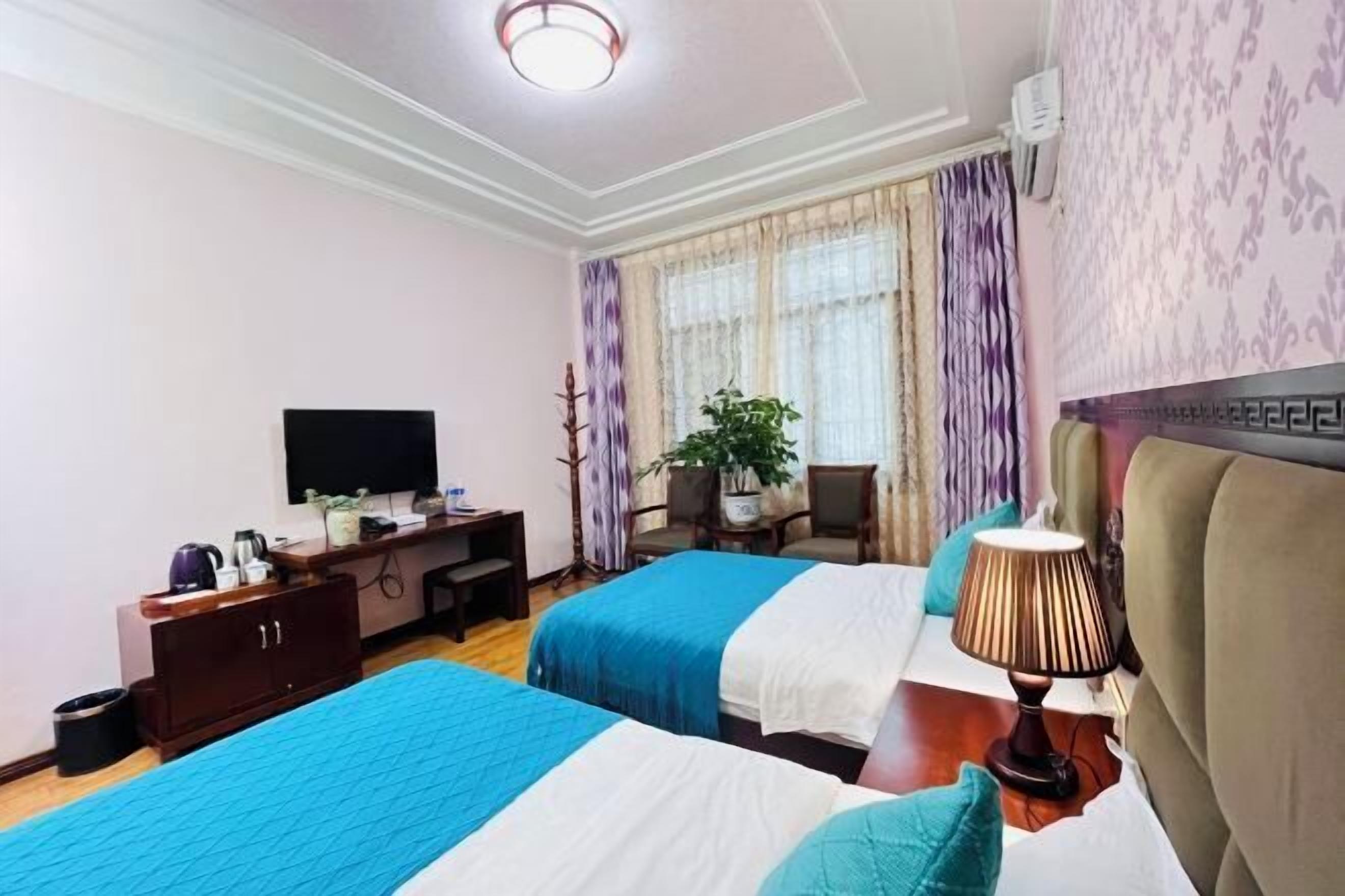 Ein Hotelzimmer mit zwei Betten, einem Schreibtisch mit Fernseher, einer Pflanze und einem Fenster mit Vorhängen.