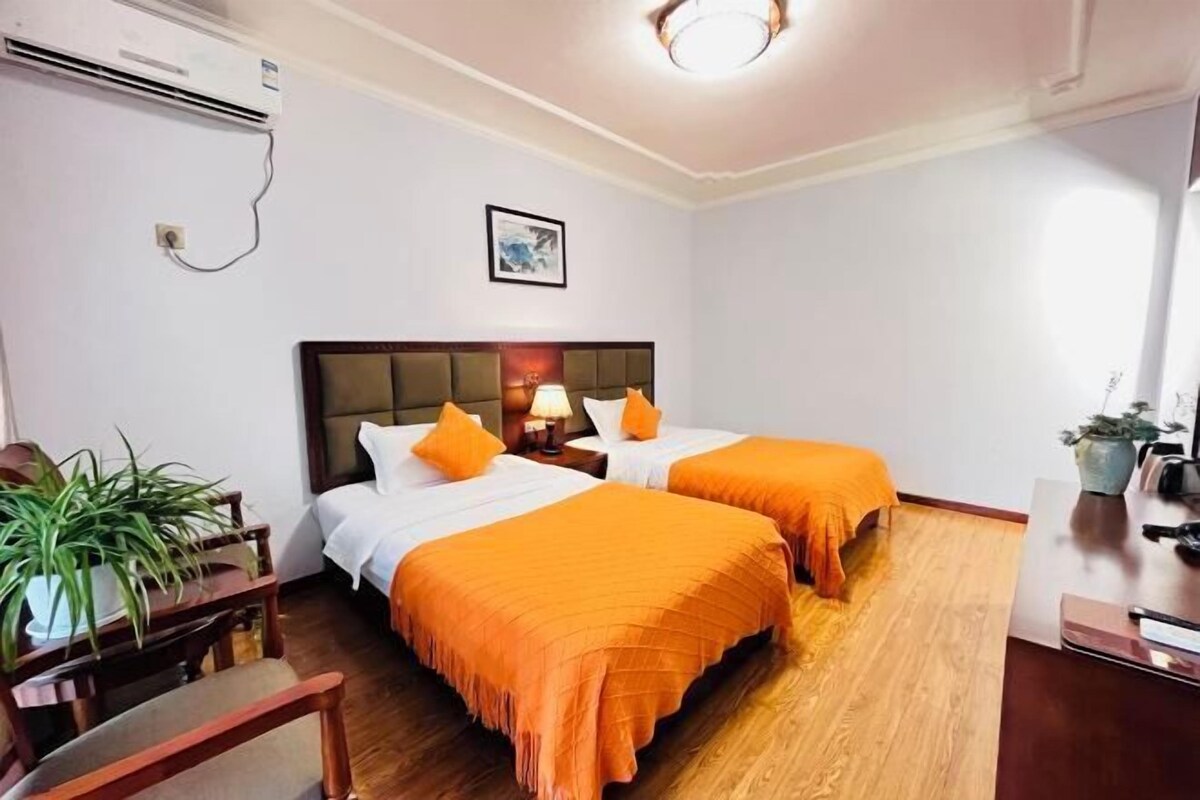 A hotel room with two beds, a desk, a chair, a potted plant, and an air conditioning unit.