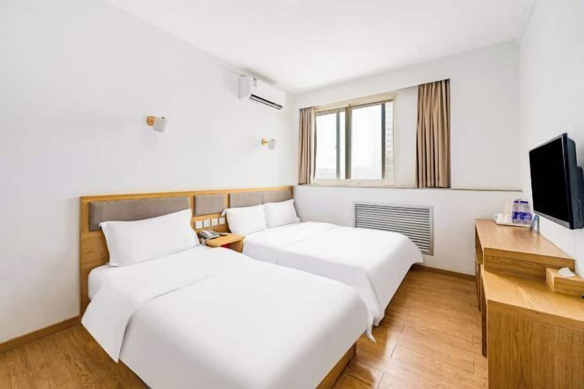 A hotel room with two beds, a TV, a desk, an air conditioner, a window with curtains, and a wall-mounted light.